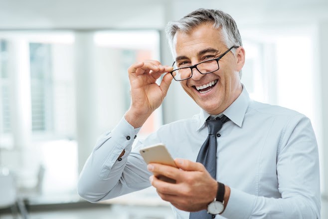 Image of a man grabbing his glasses while smiling and looking at his cell phone.