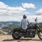 Image of a man sitting on a motorcycle with a scenic background of mountains and lakes.