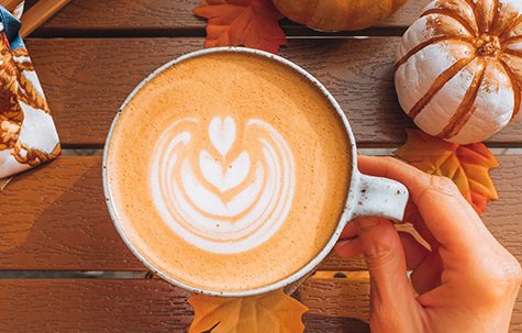 A Pumpkin Spice Latte with cream heart latte art on a wooden table with mini pumpkins surrounding.