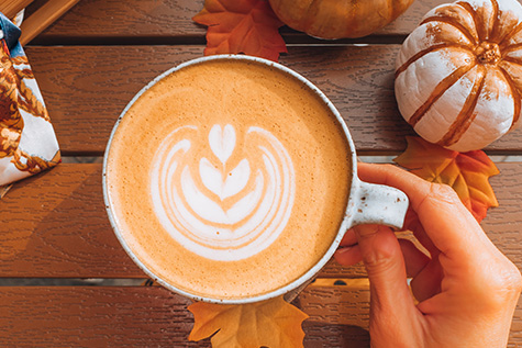 A Pumpkin Spice Latte with cream heart latte art on a wooden table with mini pumpkins surrounding.