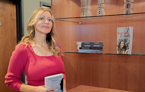 Hazel Mattice in a bright pink top holding one of her books, standing next to a display of her other books in the Unison Bank Lobby.